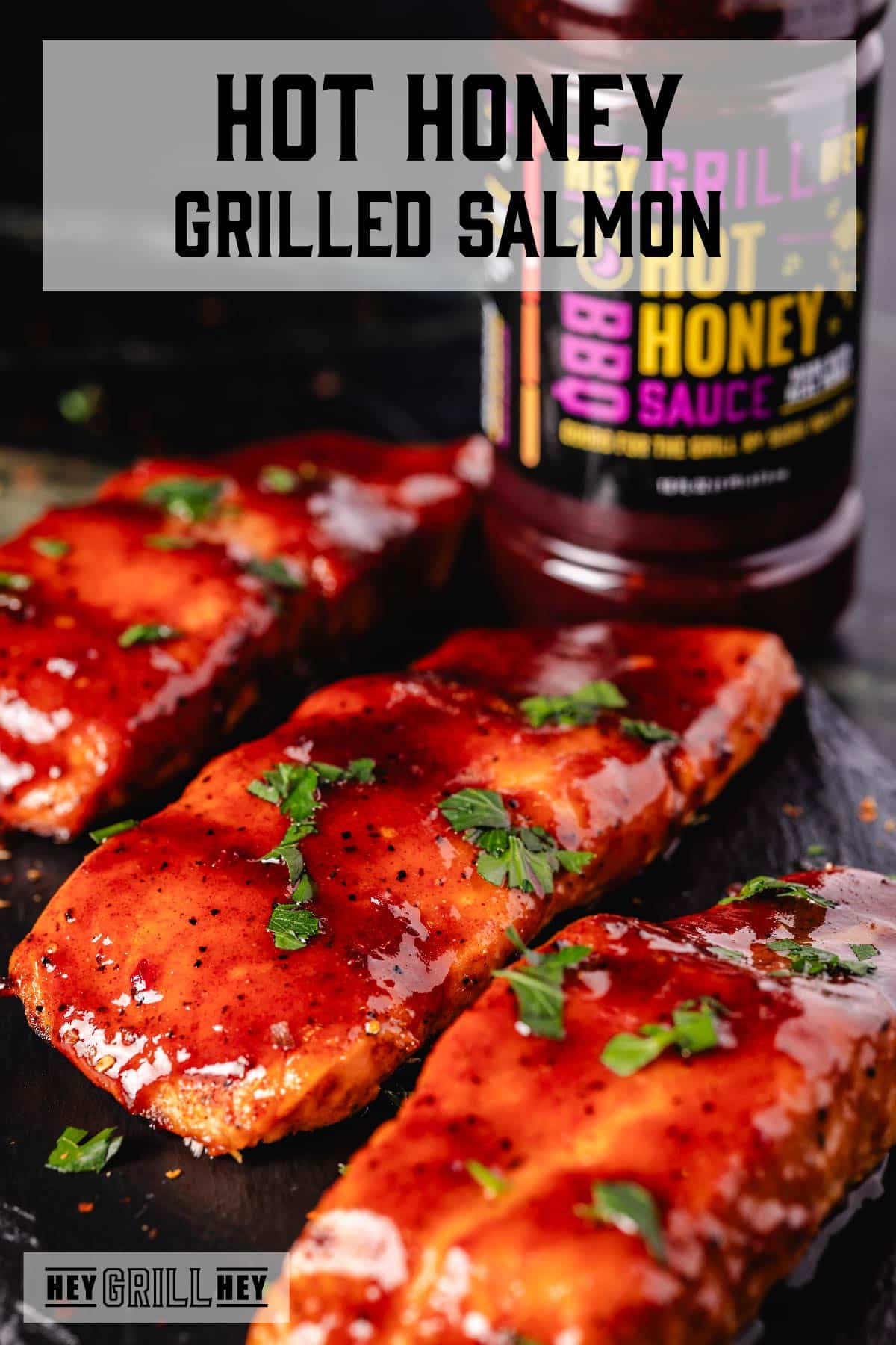 Grilled salmon fillets on a plate in front of a bottle of BBQ sauce. The text overlay reads "Hot Honey Grilled Salmon" at the top, and "Hey Grill Hey" at the bottom.