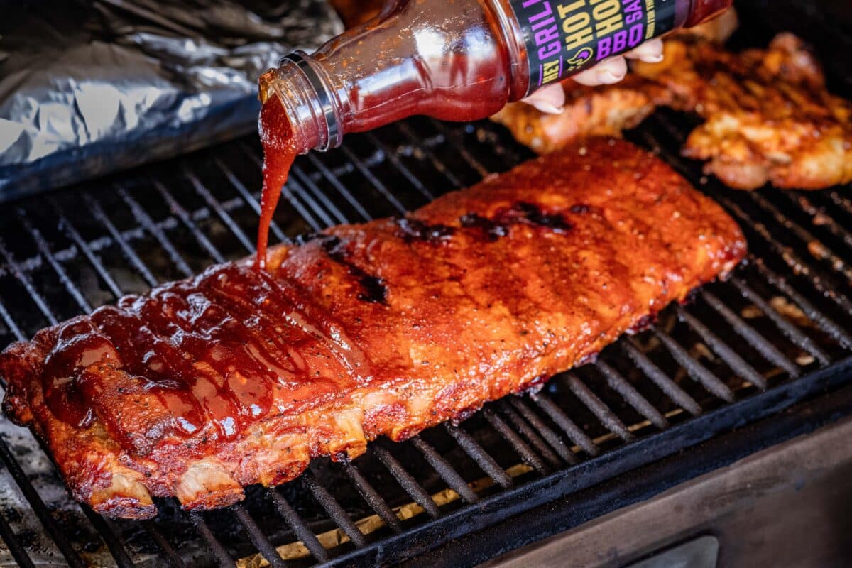 A rack of ribs on the smoker being drizzled with BBQ sauce.