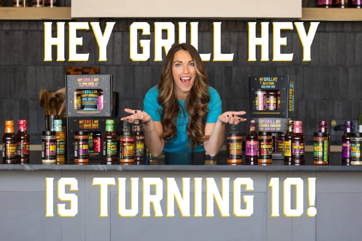 Susie from Hey Grill Hey in a blue shirt leaning on a kitchen counter, displaying several bottles and kits of BBQ sauces and rubs. The text overlay reads "Hey Grill Hey is turning 10!"