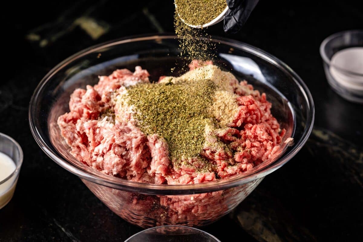 A clear glass bowl full of raw ground beef with a hand above pouring on seasoning from a small cup.