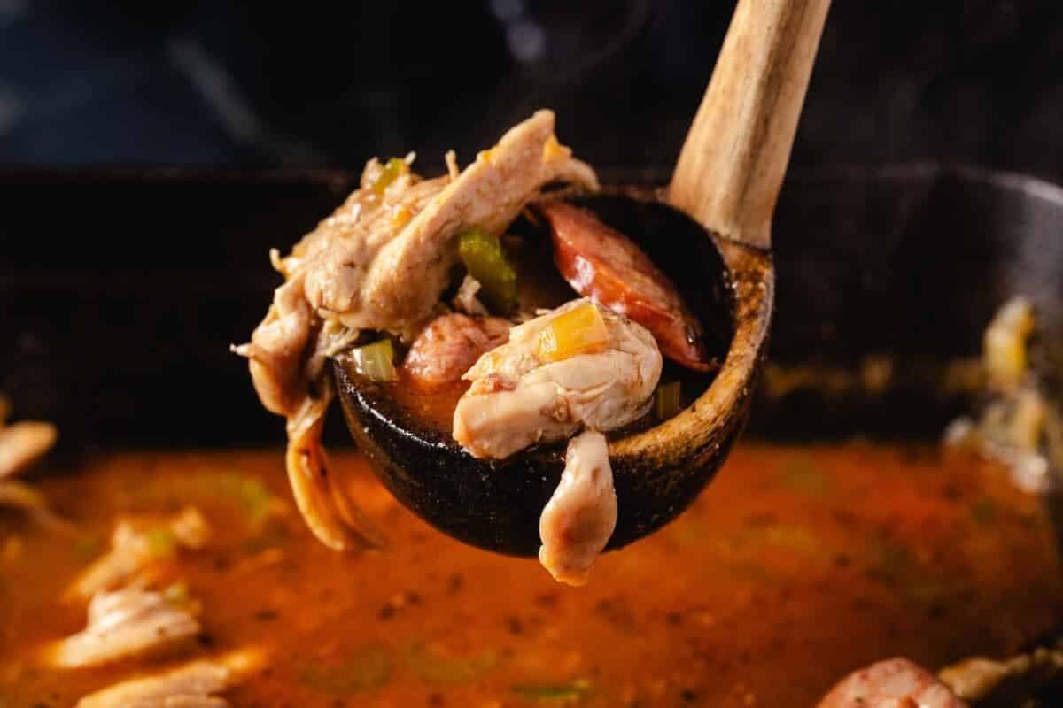 A wooden ladle holding a scoop of gumbo above a pan of it.