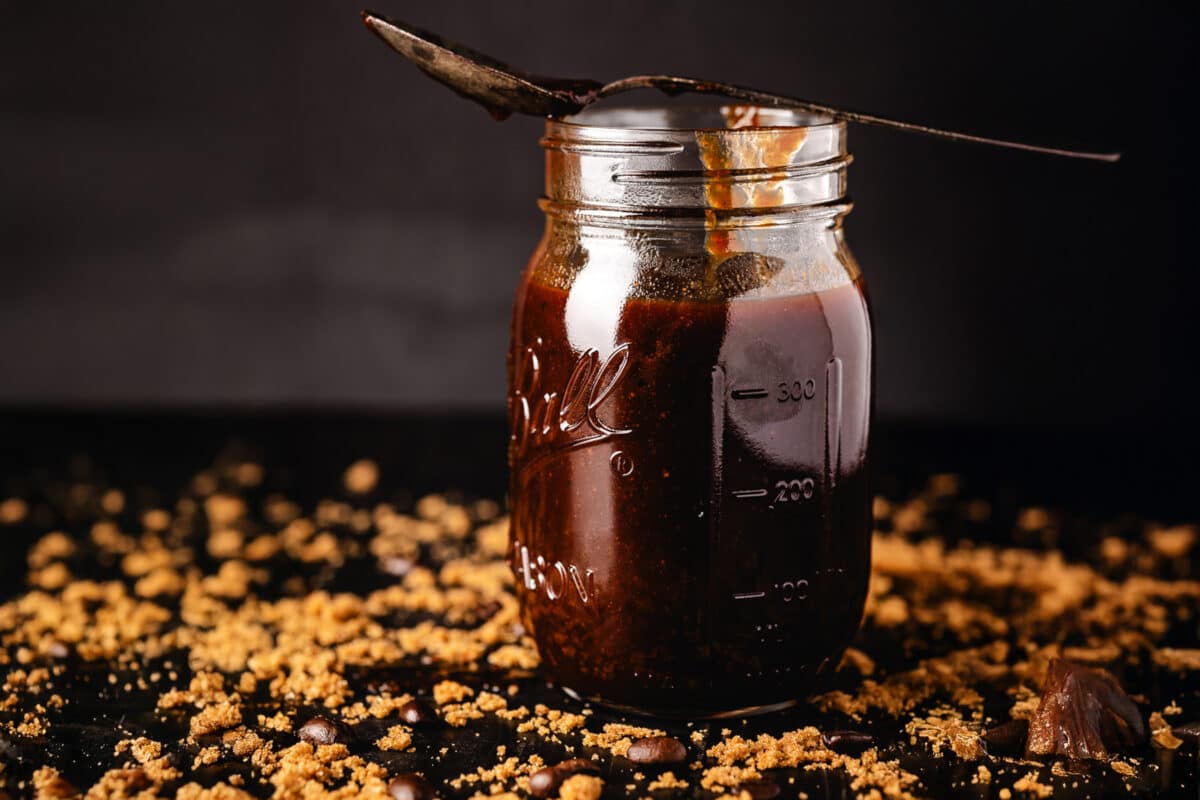 A jar of sauce with a spoon on it surrounded by loose coffee grounds.