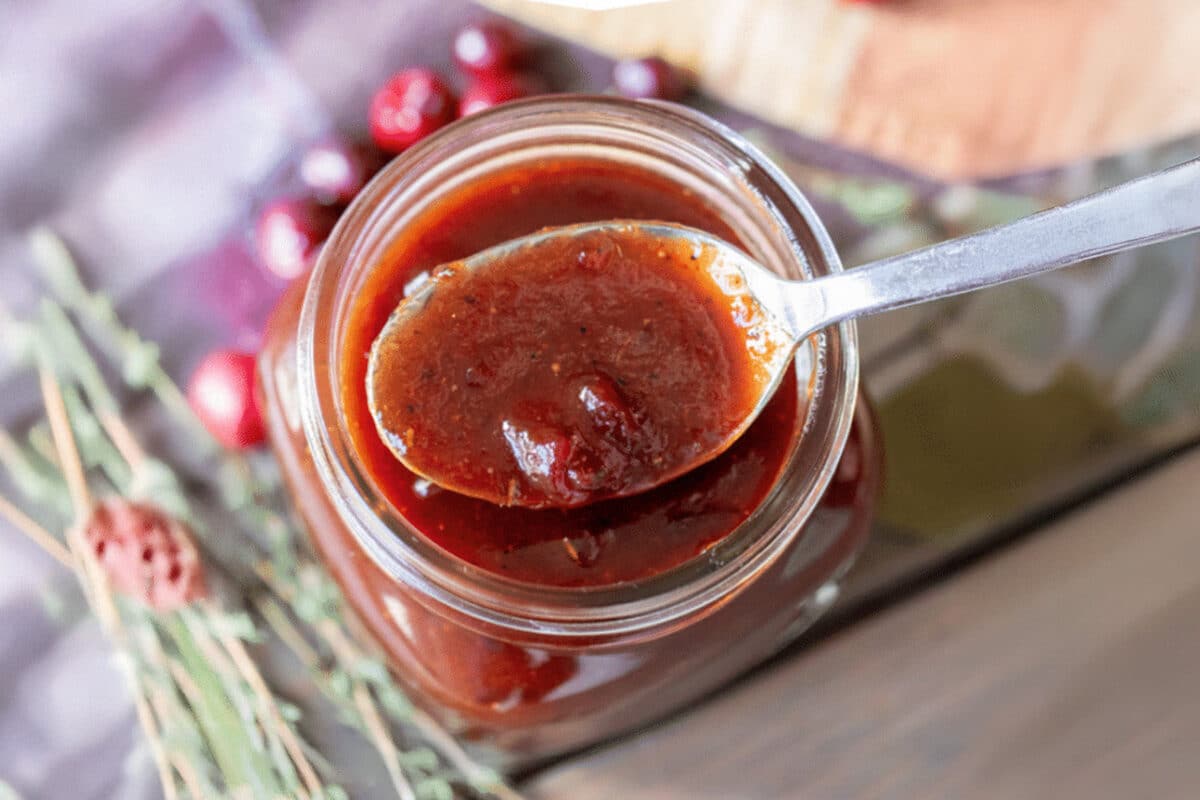 A jar of sauce with a spoon resting on it, next to fresh cranberries.