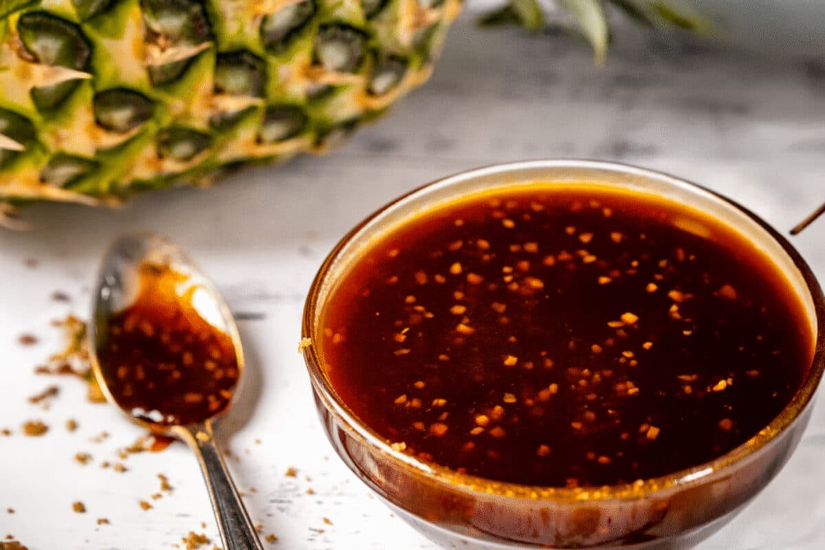 A clear bowl and spoon with BBQ sauces next to a pineapple on laying on its side.