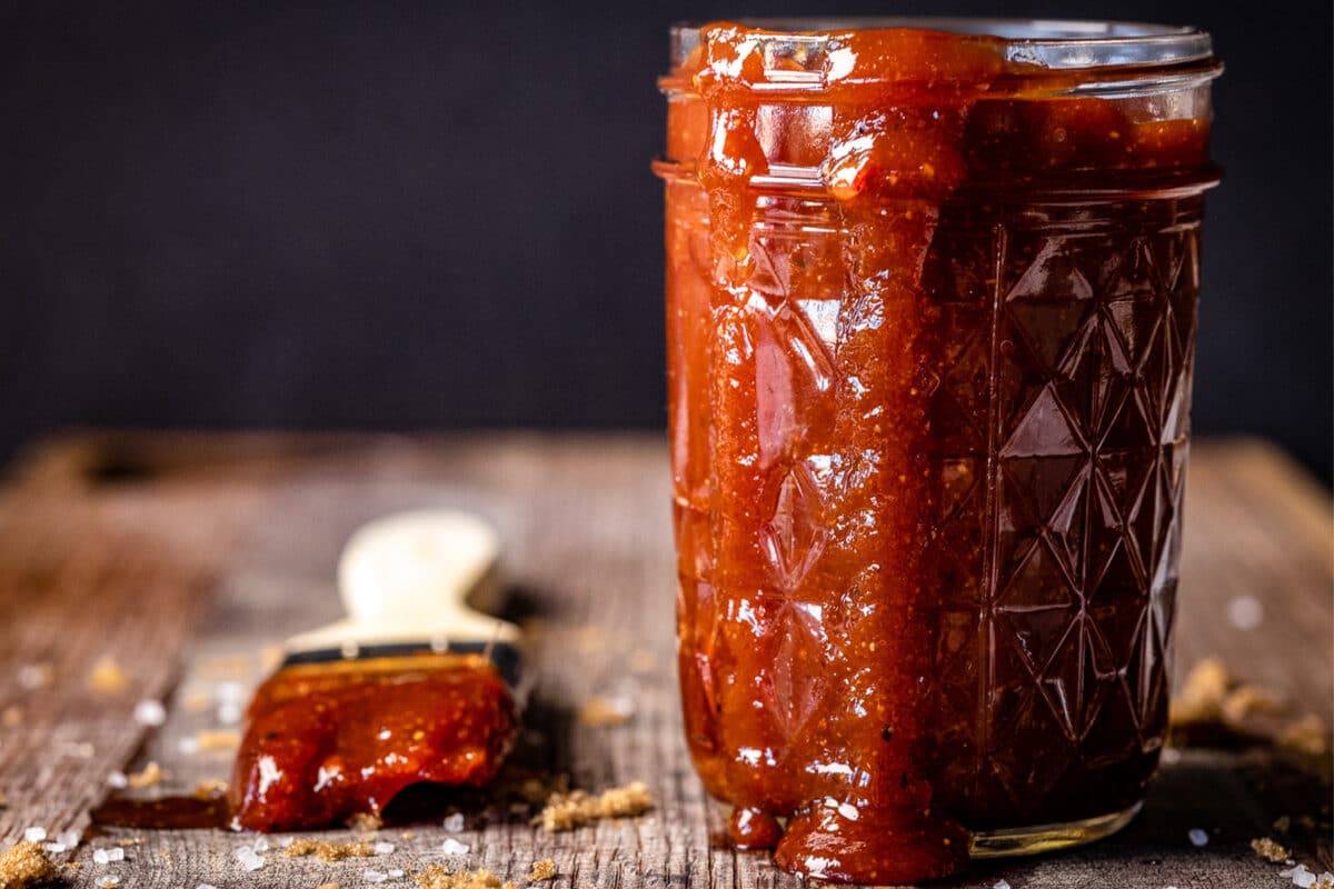 A clear glass jar of sauce with some running down the side, and a basting brush, both on a wooden table.