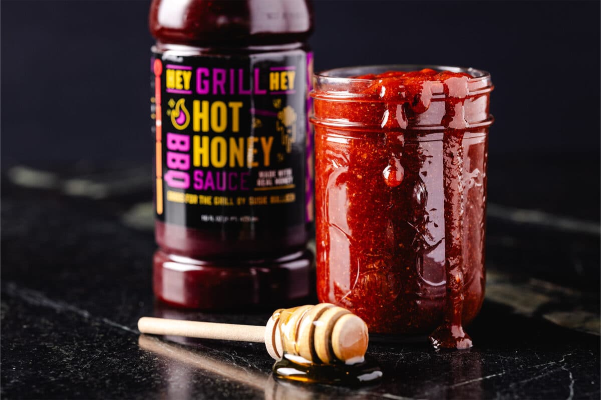 A glass jar filled and bottle of BBQ sauces next to  a honey dipper.