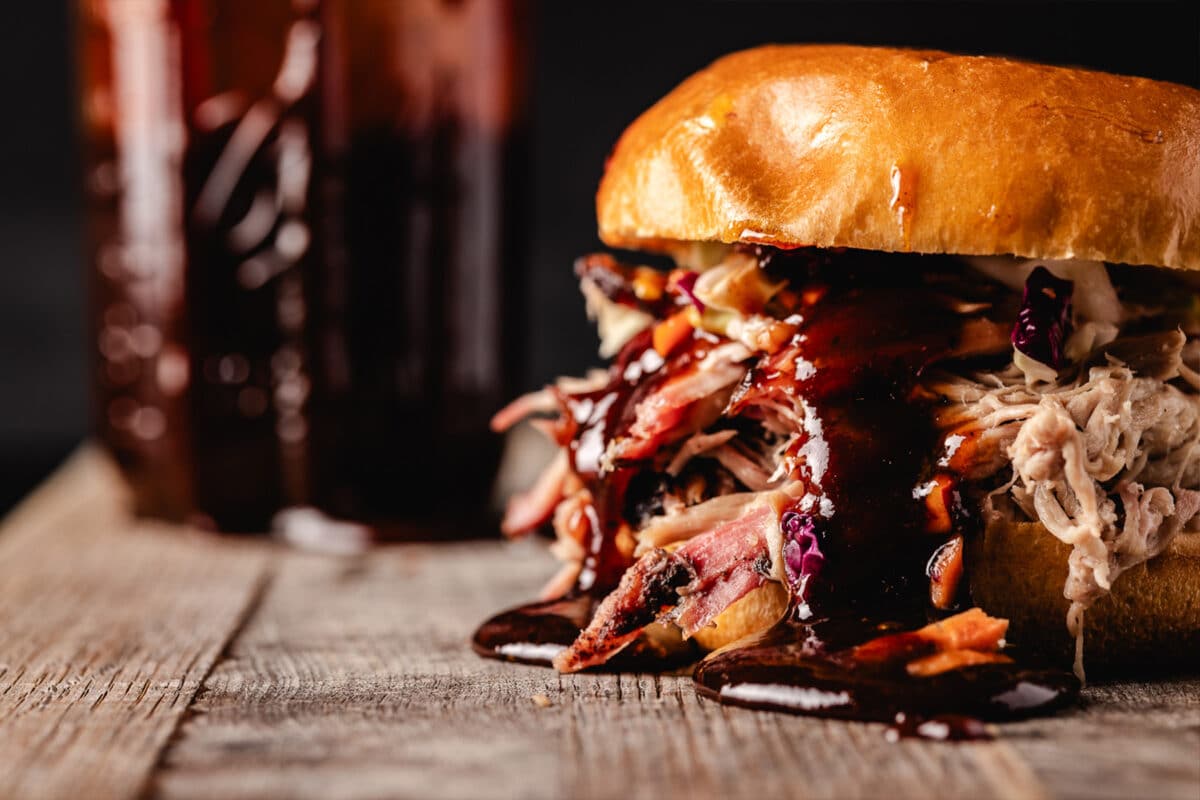 A pulled pork sandwich dripping with BBQ sauce next to a jar of the sauce.