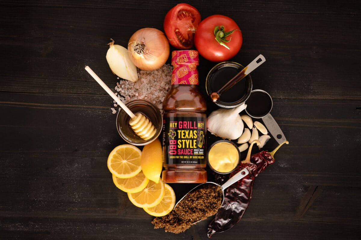 A bottle and cup of Texas BBQ sauces laying down on a surface, surrounded by the raw ingredients for it.