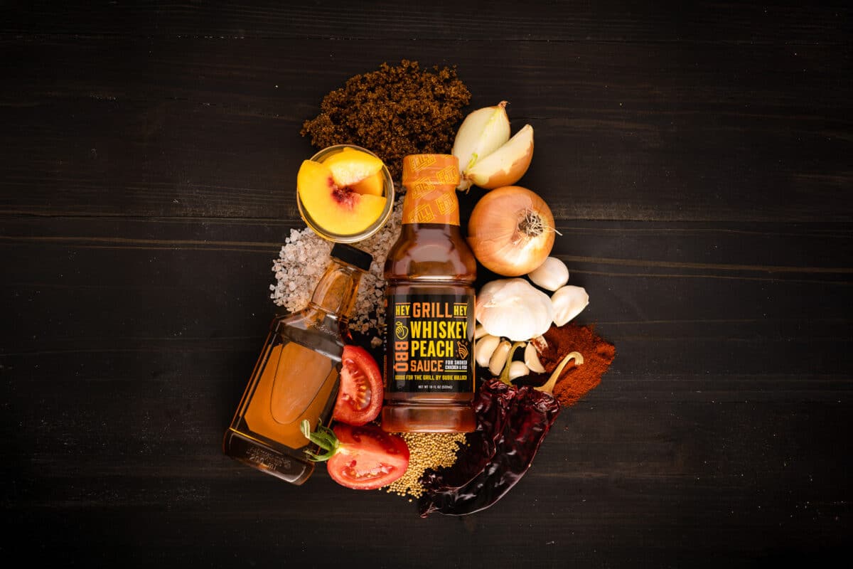 A bottle of Whiskey Peach Sauce on a table surrounded by the raw ingredients for the sauce.