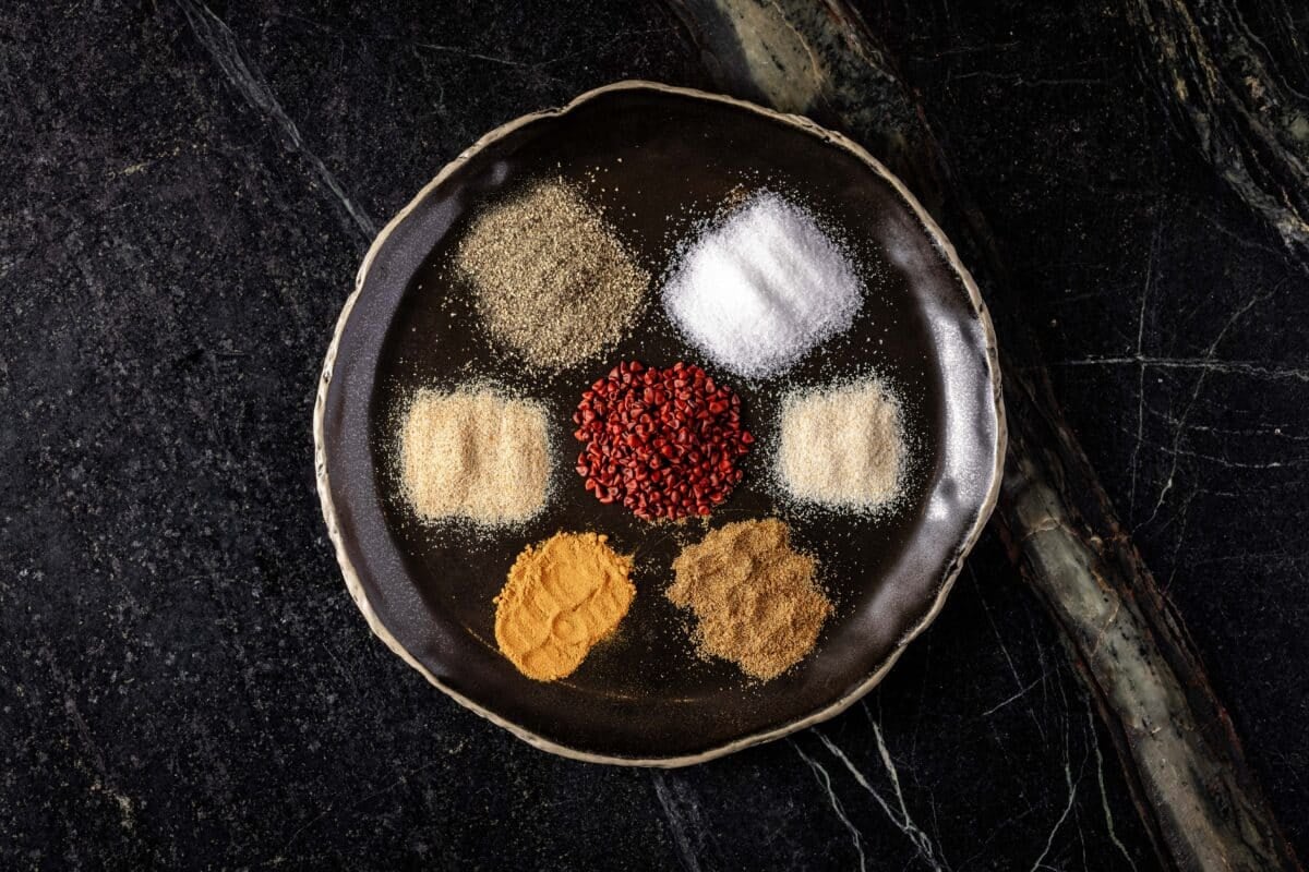 Six small piles of spices on a black plate sitting on a marble surface.