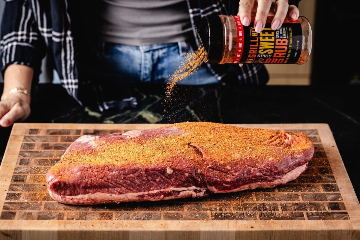 A brisket flat lying on a wooden cutting board as a hand sprinkles a bottle of Spicy Sweet Rub onto it.