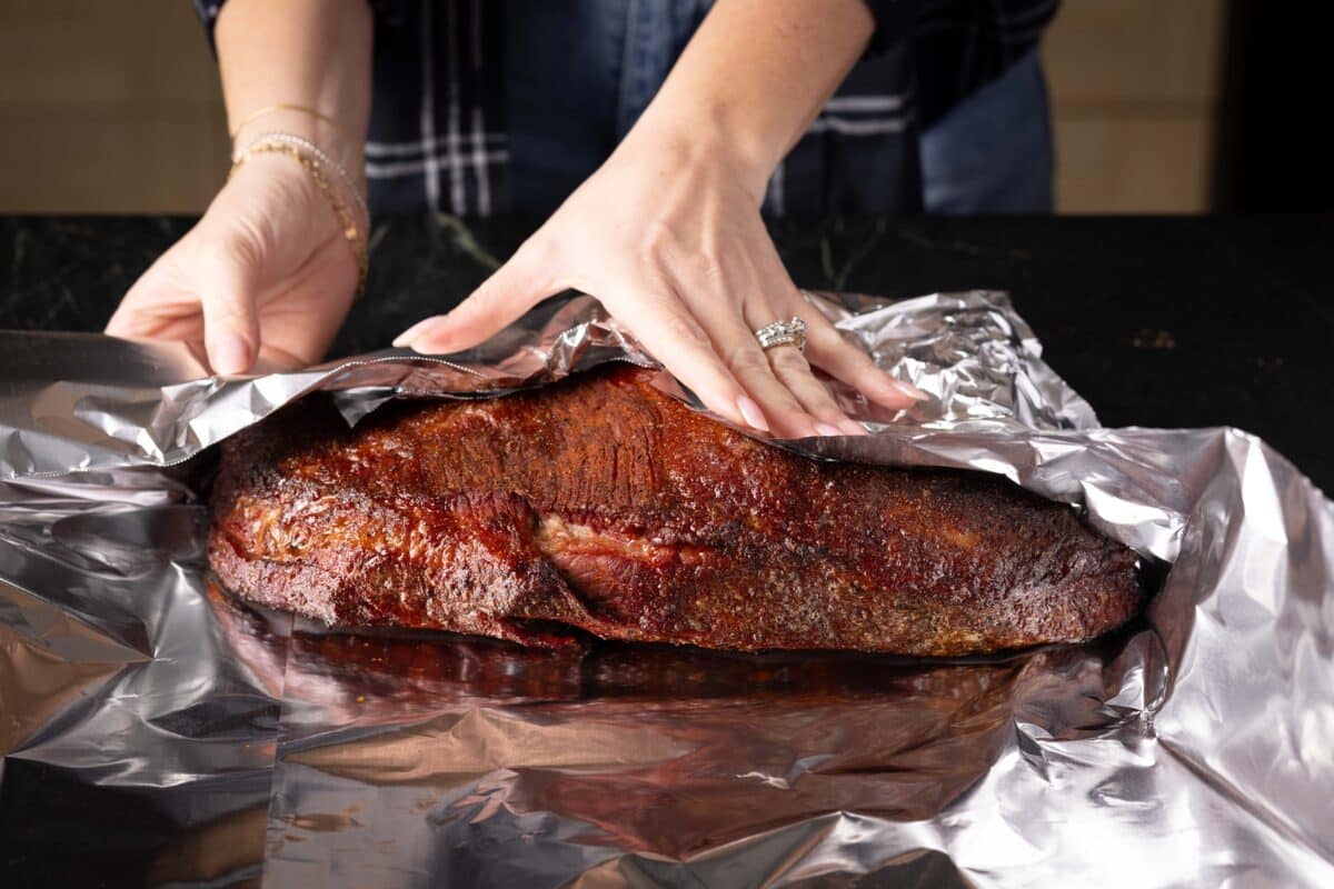 Hands wrapping a brisket flat in aluminum foil.