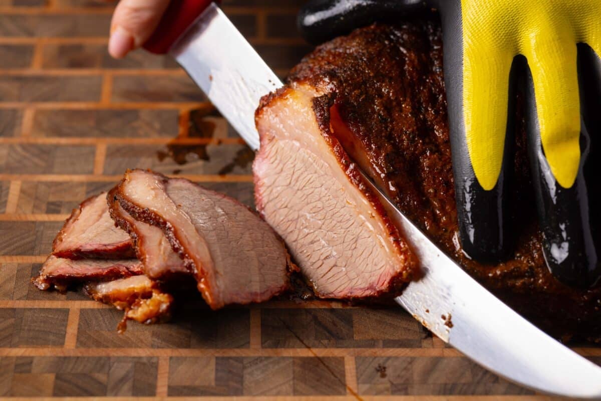 A gloved hand holding a brisket in place as the other hand slices the brisket with a knife.