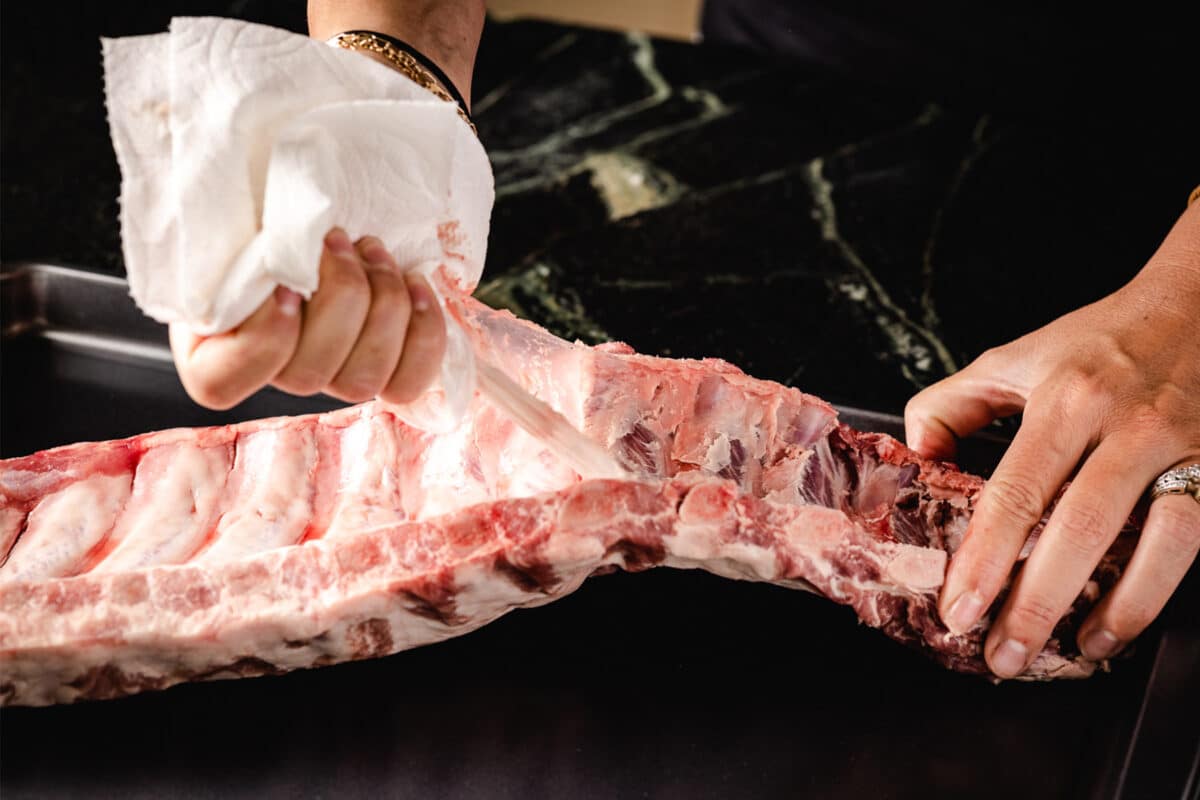 A raw rack of ribs lying on a cutting board with the membrane being pulled off.