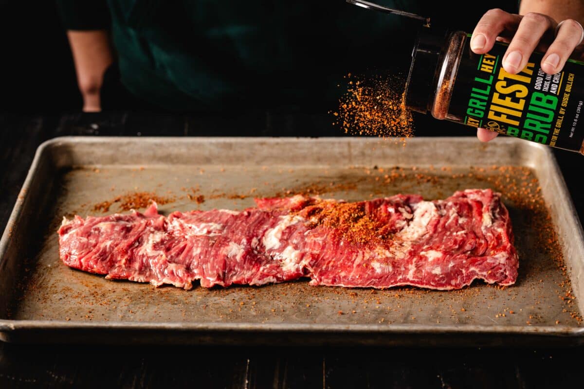 Raw flap steak on a metal baking sheet as a hand above sprinkles on Hey Grill Hey Fiesta Rub.