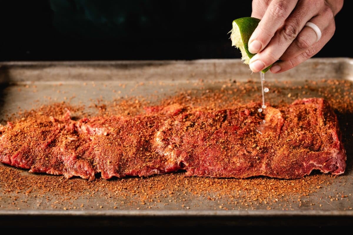 A hand holding a lime wedge, squeezing juice from it onto raw steak resting on a baking sheet.