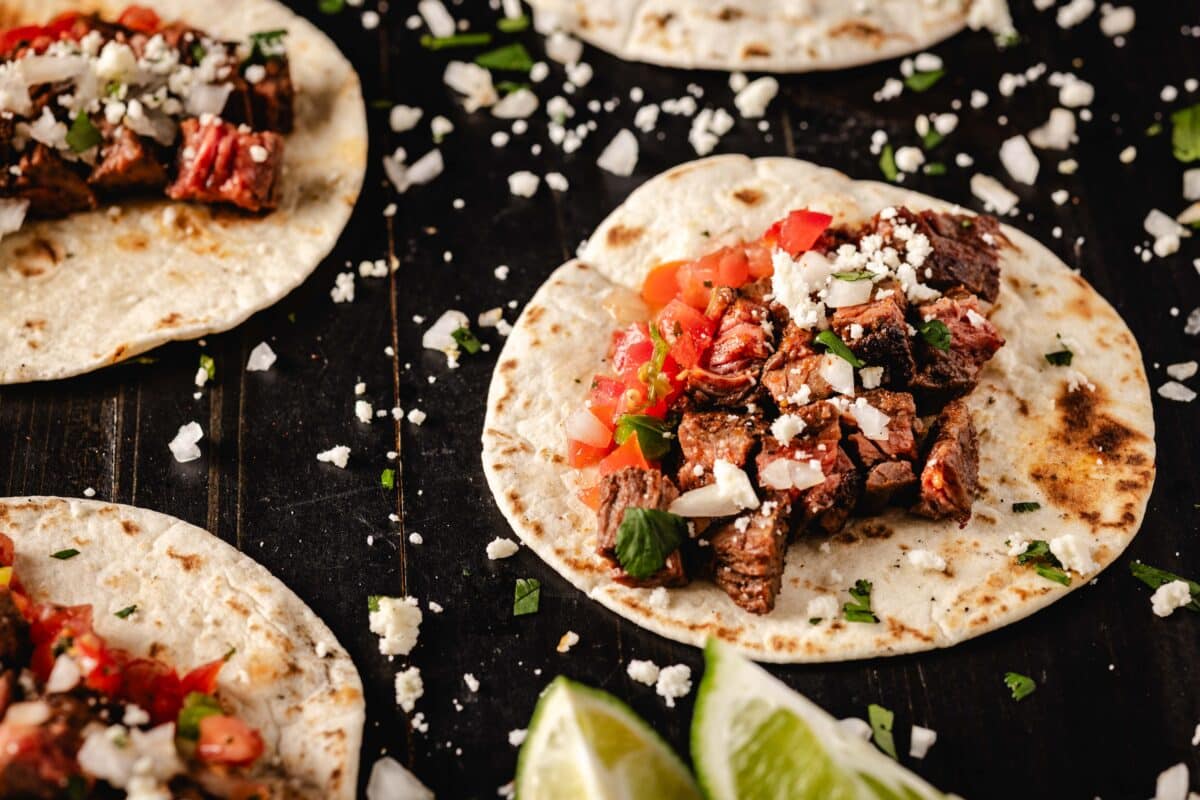 Several carne asada tacos on a black countertop which is sprinkled with shredded cheese.