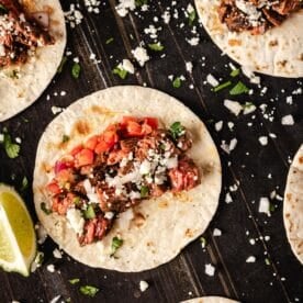 Several carne asada tacos topped with salsa on a black surface sprinkled with cheese.