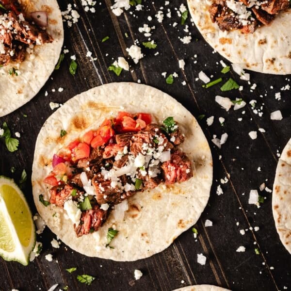 Several carne asada tacos topped with salsa on a black surface sprinkled with cheese.