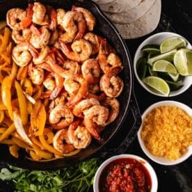 A skillet filled with cooked shrimp next to while bowls off shredded cheese, lime wedges, and salsa.