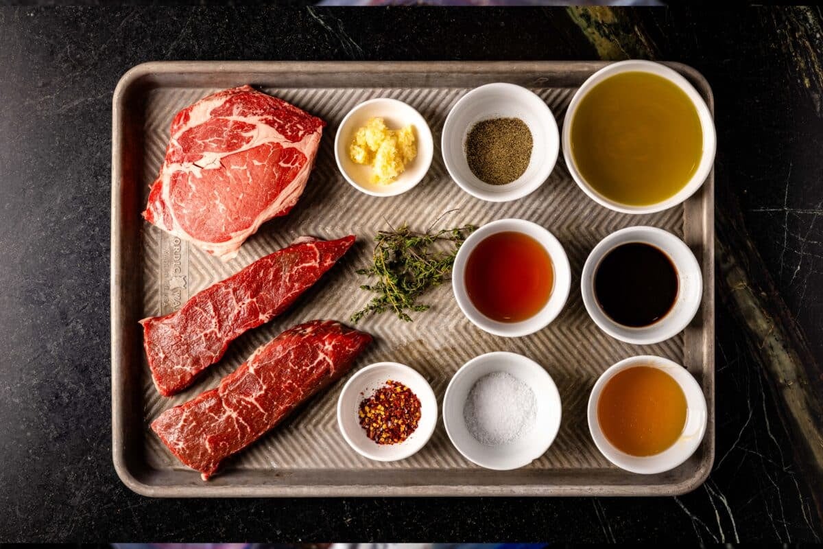 Raw cuts of beef and bowls with various ingredients arranged on a baking sheet.