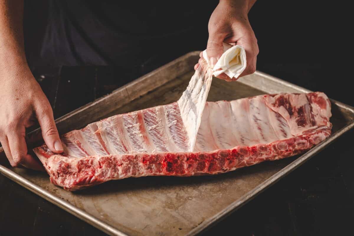 An uncooked rack of ribs on a baking sheet, as a hand peels back the membrane to remove it.