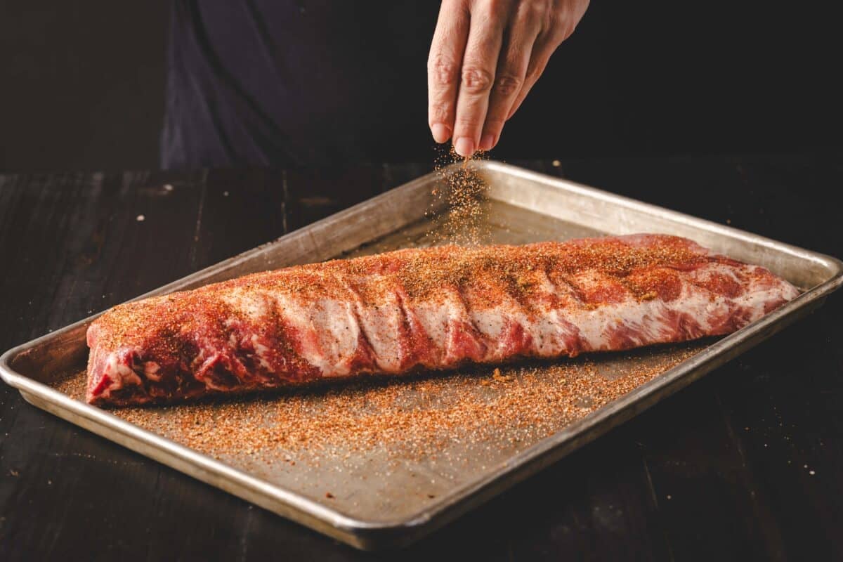 A hand sprinkling a rub onto a rib rack on a pan below.