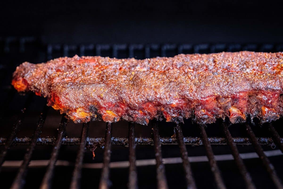 A seasoned rack of baby backs on the grates of a smoker with the bone starting to show.