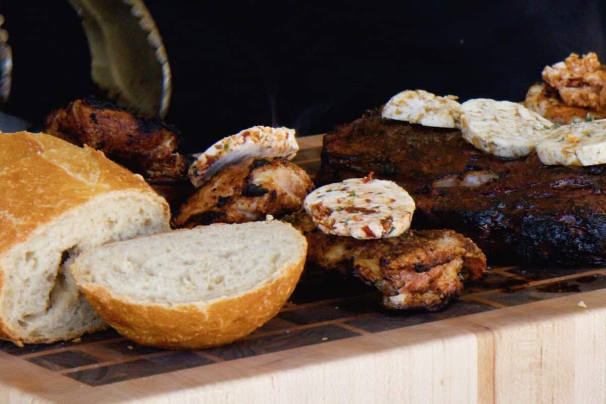 Grilled chicken resting on a cutting board next to a partially-sliced loaf of bread and a cooked steak.