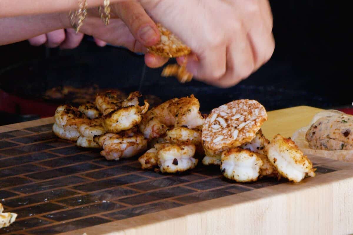 Grilled shrimp resting on a cutting board as hands places a seasoned topping on.