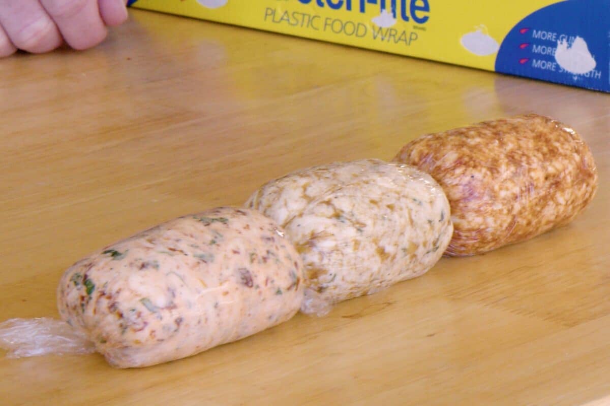 Various compound butters wrapped in shrink wrap on a wooden cutting board.