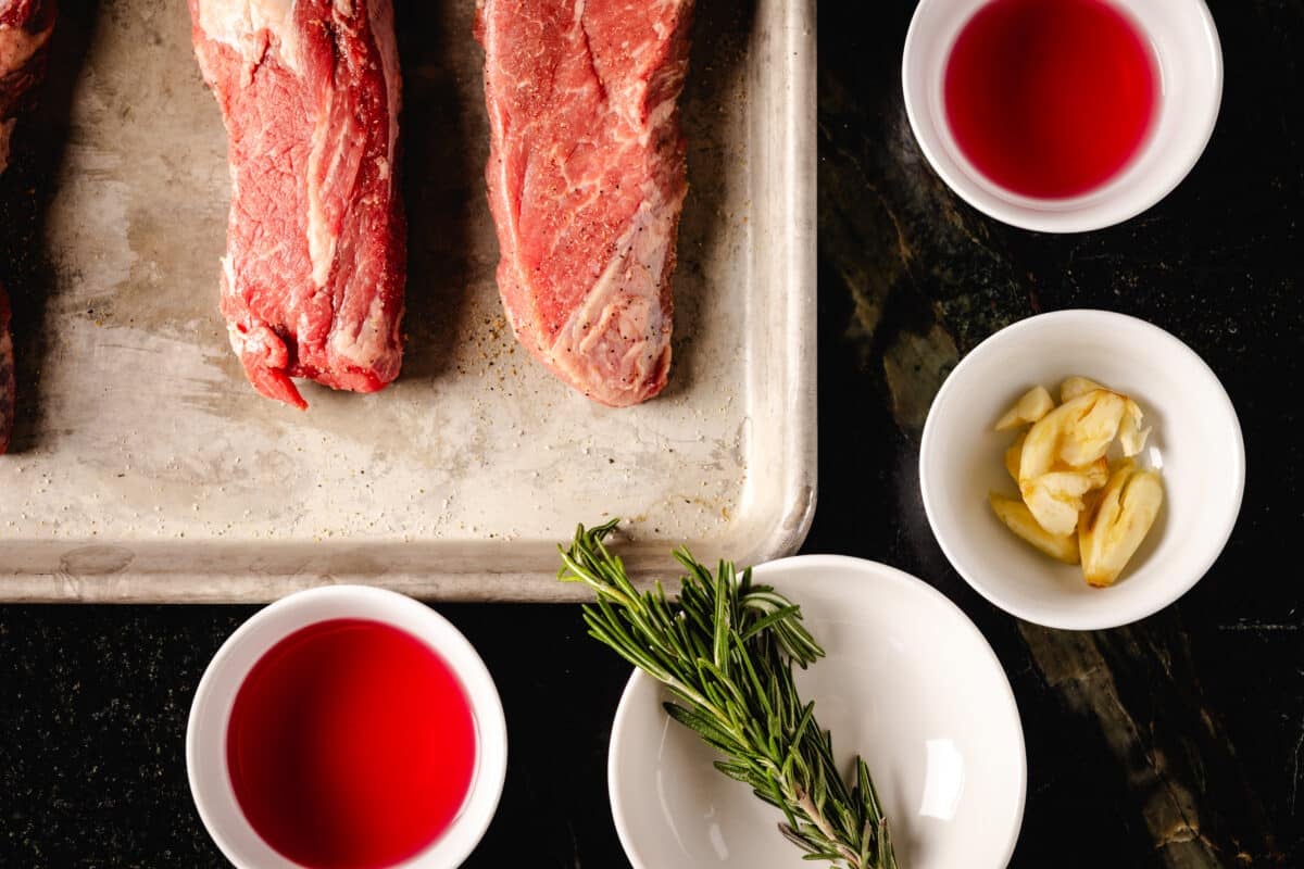 Ingredients for a steak marinade in white bowls next to a baking sheet topped with raw steaks.