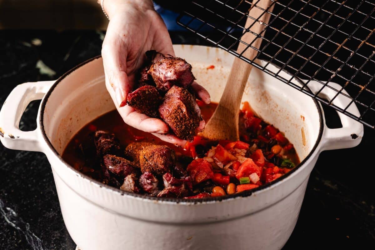 A hand adding cubes of smoked beef from a wire rack into a white pot of chili below.