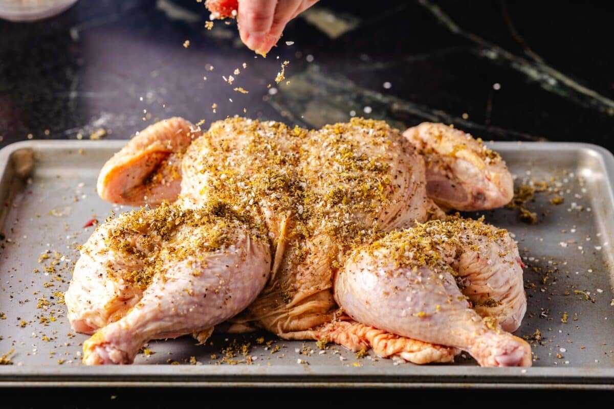 A spatchcock chicken on a baking sheet being sprinkled with seasoning from a hand above.