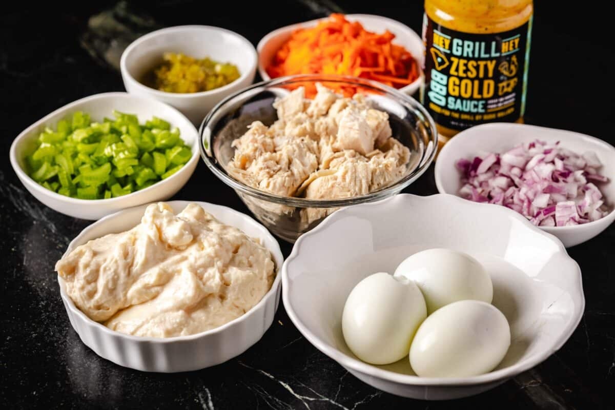 A metal bowl filled with tuna, surrounded by white glass bowls with eggs, and chopped veggies.