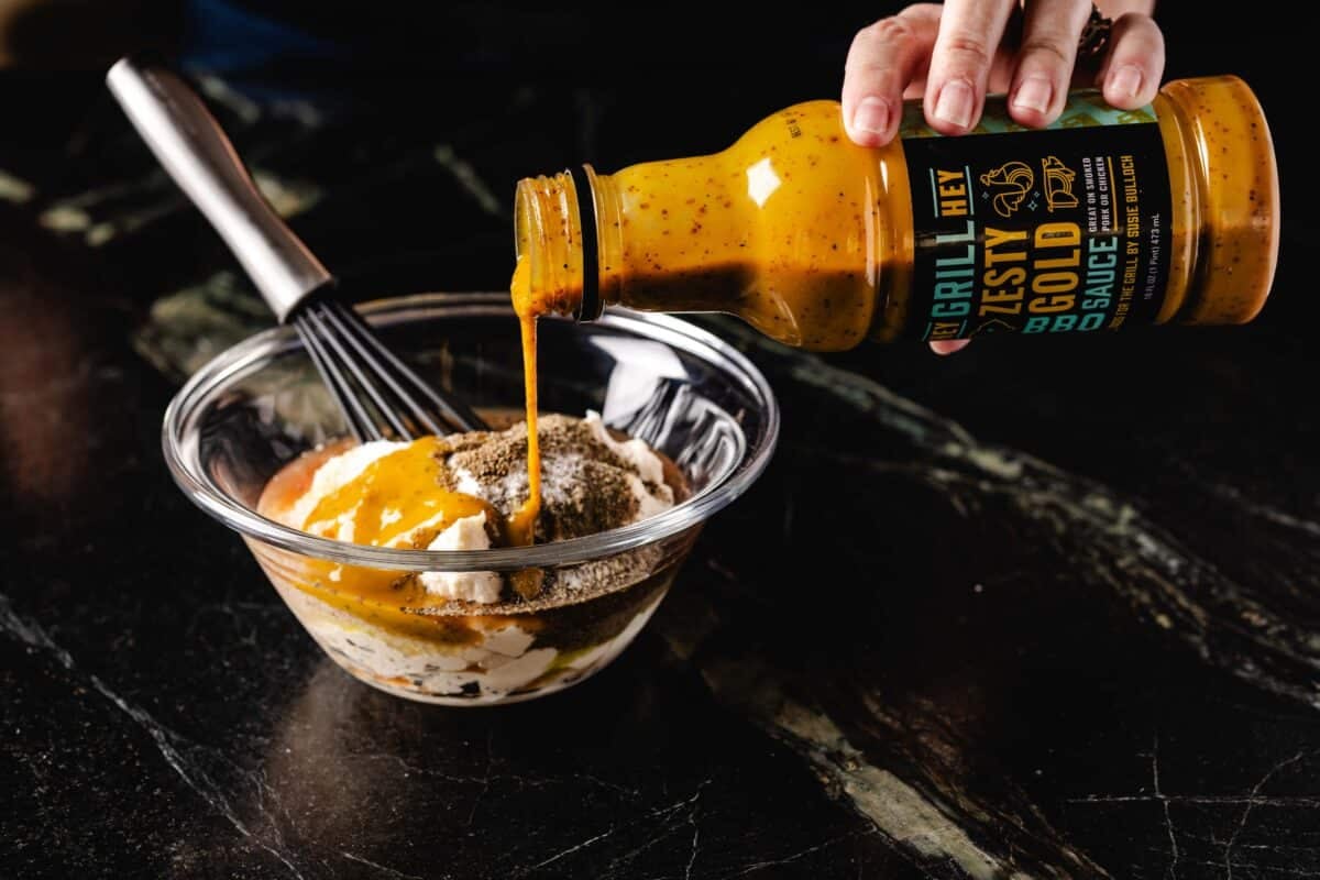 A bottle of Hey Grill Hey Zesty Gold BBQ Sauce pouring into a bowl of ingredients.