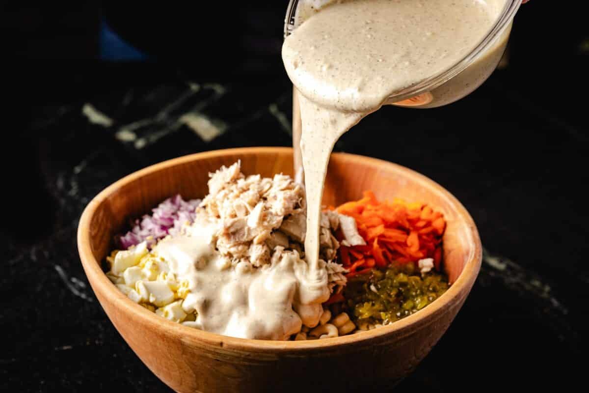 A bowl of dressing being poured into a bowl or salad ingredients including tuna and macaroni noodles.