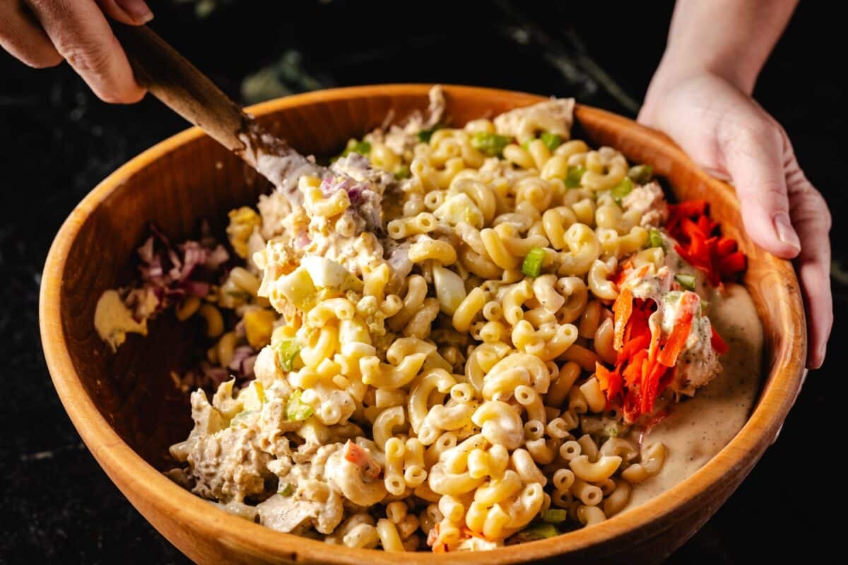 A hand using a wooden spoon to stir a macaroni salad in a wooden bowl.