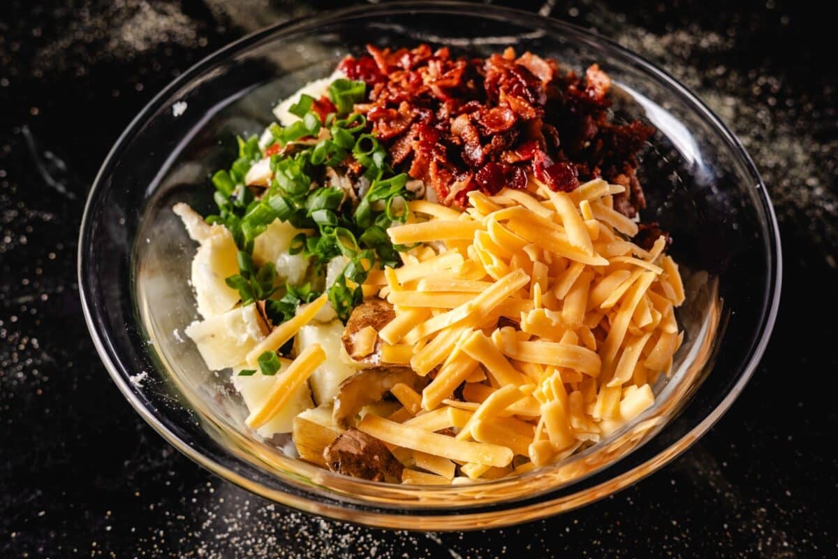 Crumbled bacon, sliced green onions, shredded cheese, and cubed potatoes in a clear bowl on a marble surface.