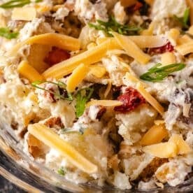 A closeup shot of a clear bowl full of a potato salad topped with shredded cheese.