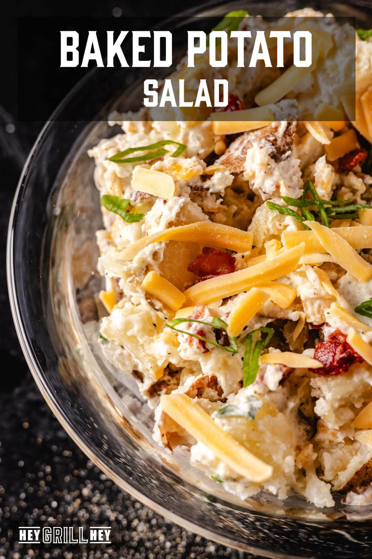 A closeup shot of a clear bowl full of a potato salad topped with shredded cheese. The text overlay reads "Baked Potato Salad" at the top, and "Hey Grill Hey" at the bottom.