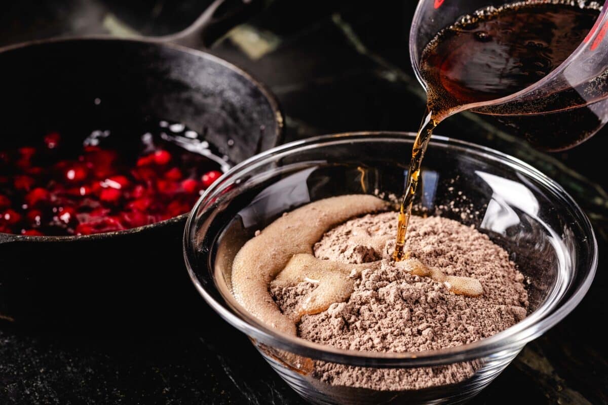 A plastic measuring cup pouring a brown liquid into a clear bowl filled with chocolate cake mix.