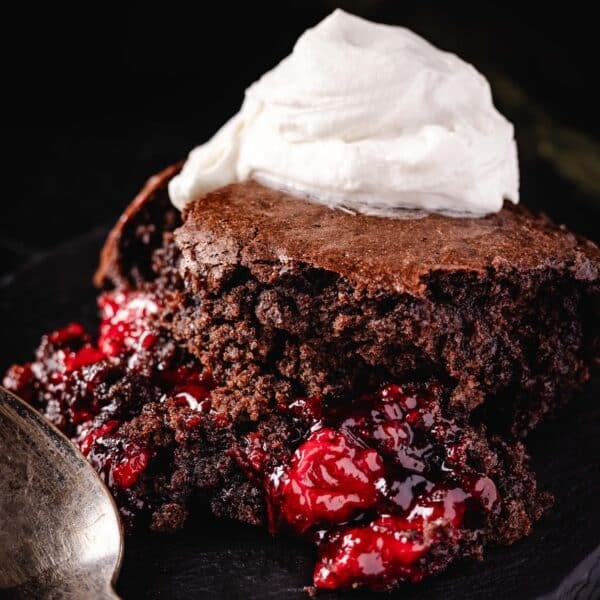 A slice of chocolate cherry cobbler topped with whipped cream on a black plate next to a spoon.