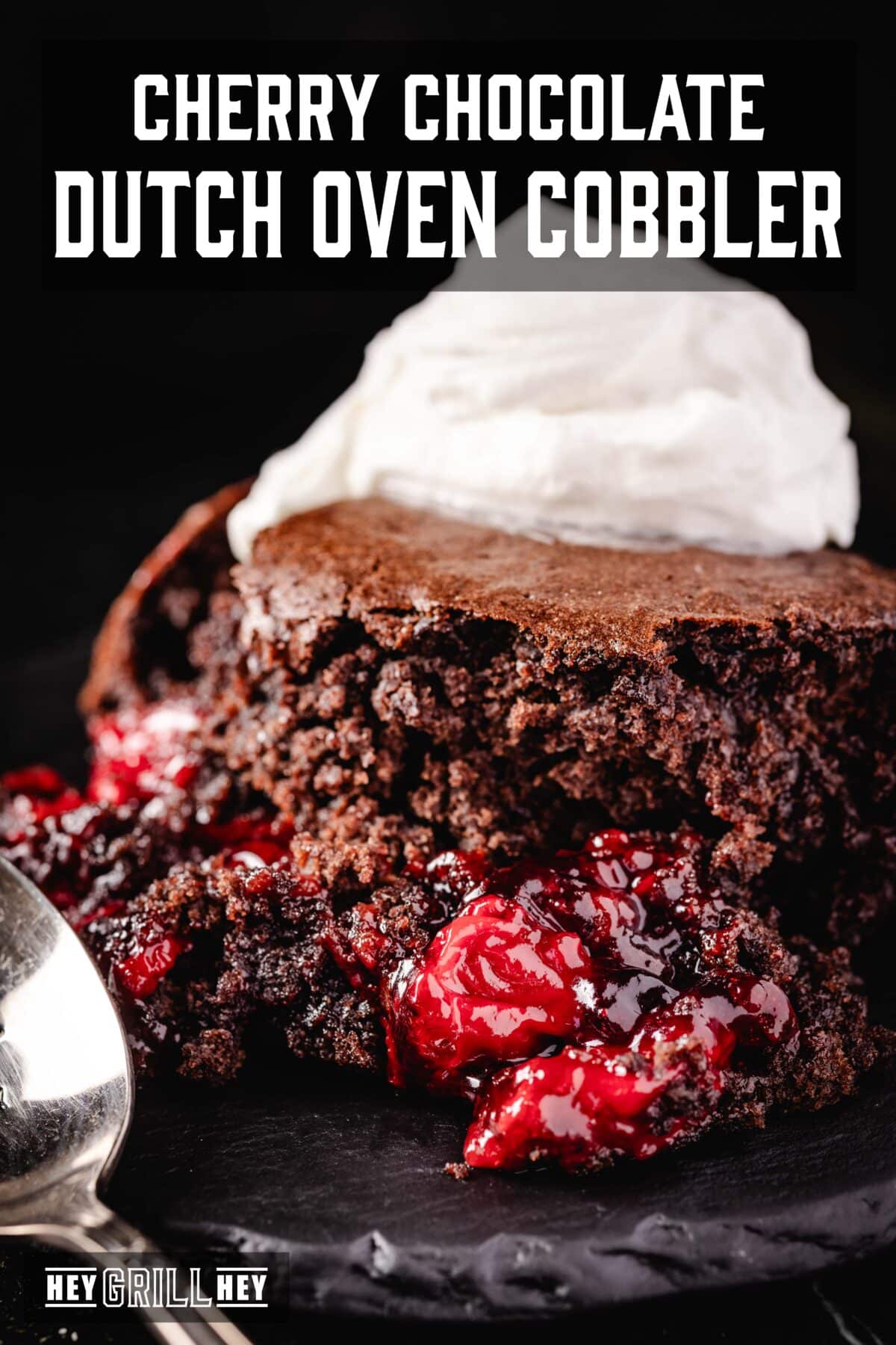 A slice of chocolate cherry cobbler topped with whipped cream on a plate next to a spoon. The text overlay reads "Cherry Chocolate Dutch Oven Cobbler" at the top, and "Hey Grill Hey at the bottom.