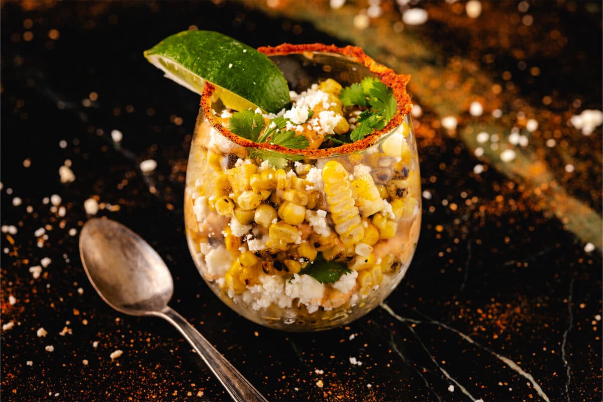 A glass filled with esquites garnished with lime, cilantro, and cotija cheese on a black table with a serving spoon.