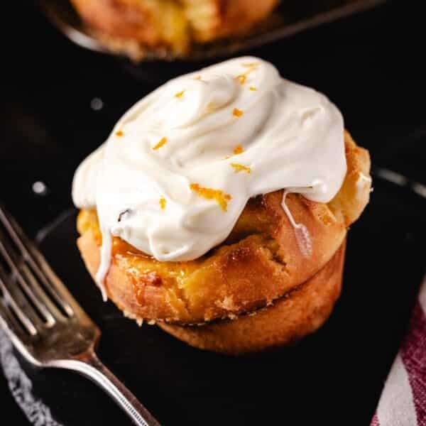 A frosted orange roll on a black plate next to a fork.