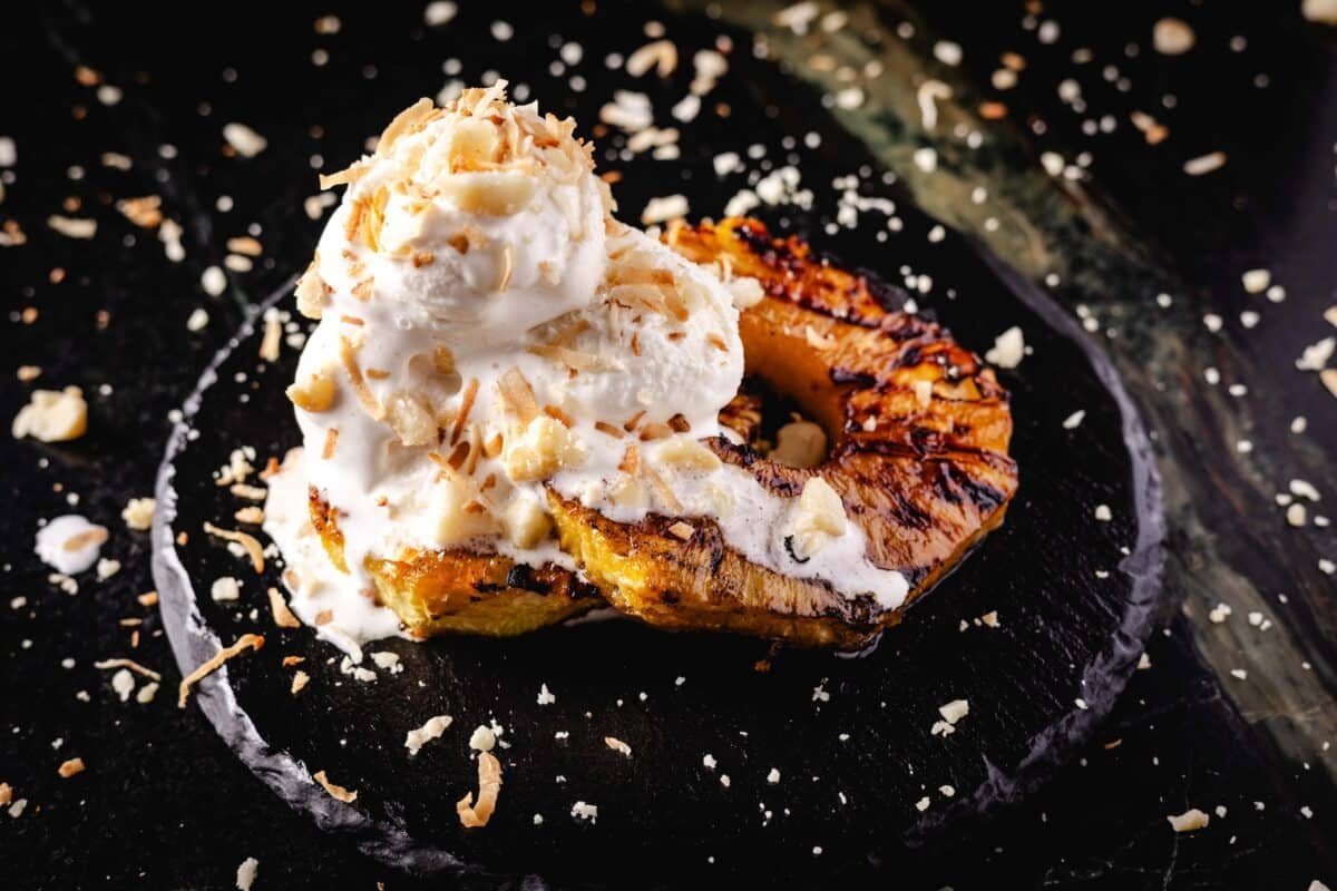 A grilled fruit dessert topped with coconut ice cream and a nut crumble on a black plate.