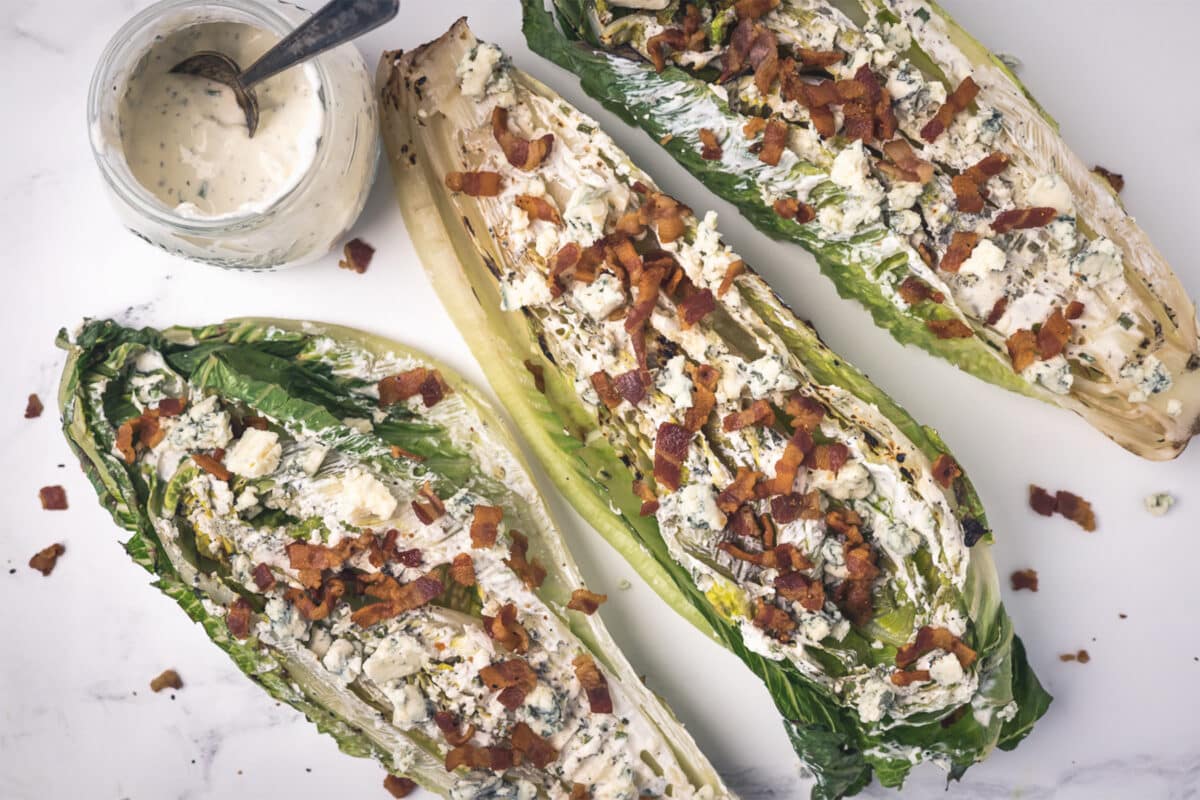 Three servings of grilled romaine lettuce topped with bacon and blue cheese crumbles on a white serving dish with a side of ranch.