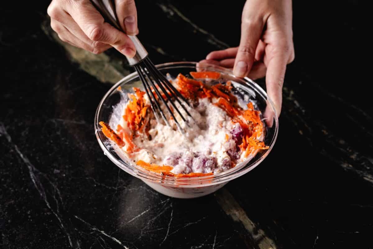 One hand holding a bowl of dressing in place as another hand whisks shredded carrots into the dressing.