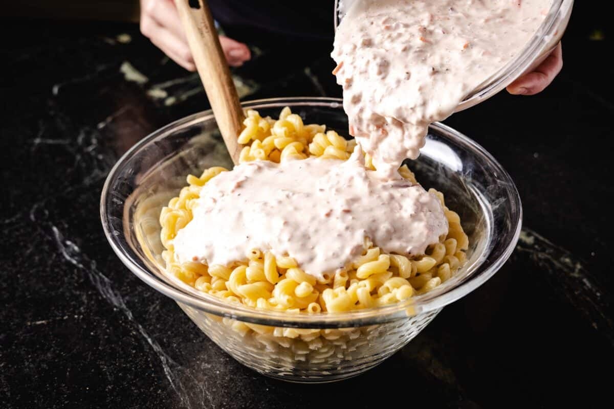 A bowl of dressing being poured into a bowl of pasta noodles.